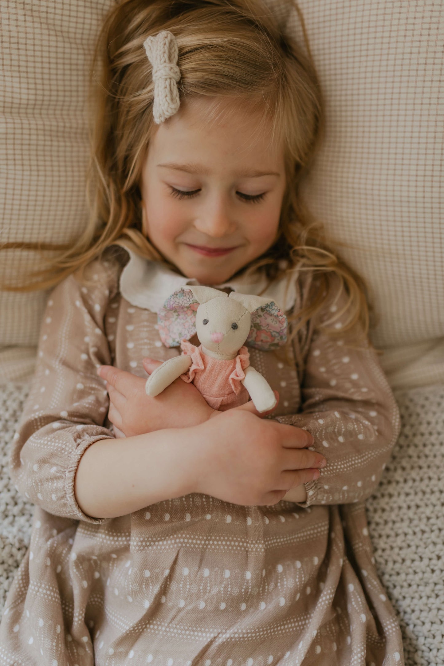 Young girl hugging the Betsy Bunny toy, wearing a patterned dress and headband, lying on a textured surface.